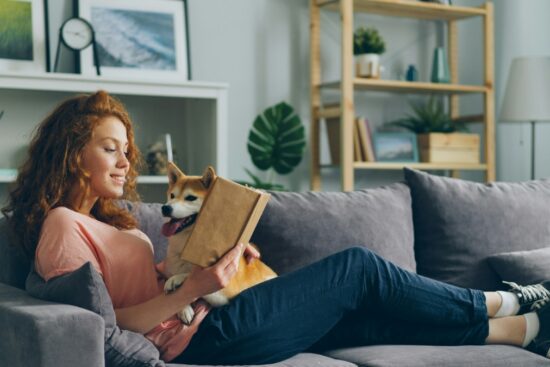 Woman relaxing on a gray sofa with a book and a happy dog in a cozy living room.