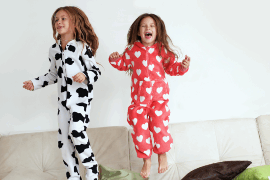 Two girls jumping on a couch wearing cozy one-piece pajamas, one in a cow print and the other in red with white hearts.
