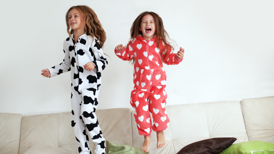 Two girls jumping on a couch wearing cozy one-piece pajamas, one in a cow print and the other in red with white hearts.