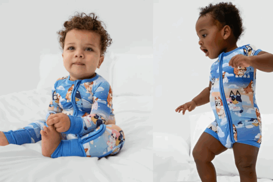Two toddlers in blue character onesies, one sitting and one standing on a white bed.
