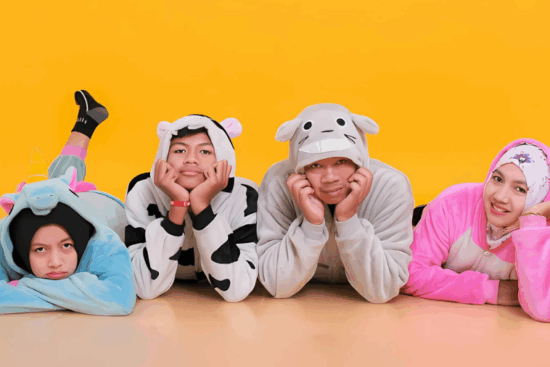 Four people wearing colorful animal-themed onesies posing playfully on the floor against a bright yellow background.
