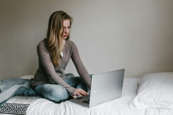 A woman sitting cross-legged on a bed using an Acer Chromebook laptop.