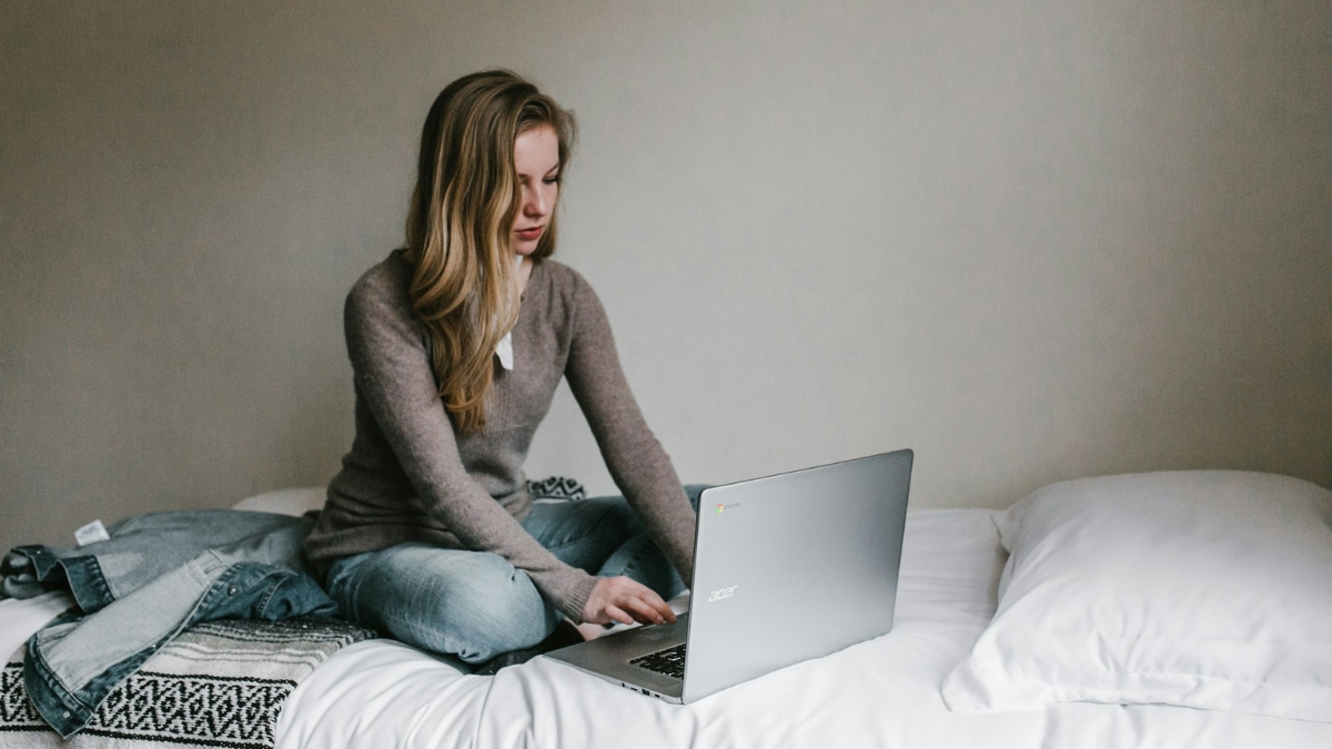 A woman sitting cross-legged on a bed using an Acer Chromebook laptop.