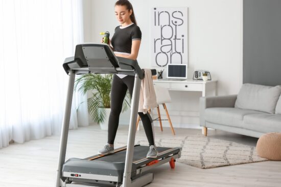 Woman using a home treadmill in a bright living room with a towel and water bottle.