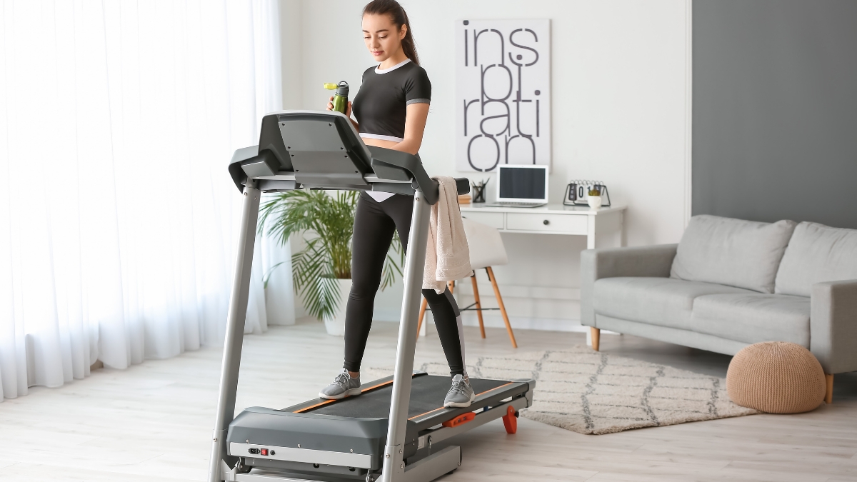 Woman using a home treadmill in a bright living room with a towel and water bottle.