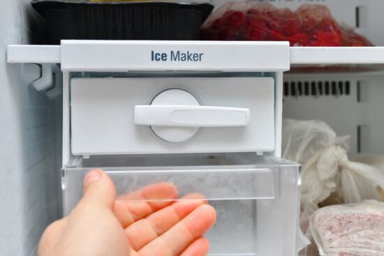 Hand opening a freezer drawer under a home ice maker with frozen food bags around it.