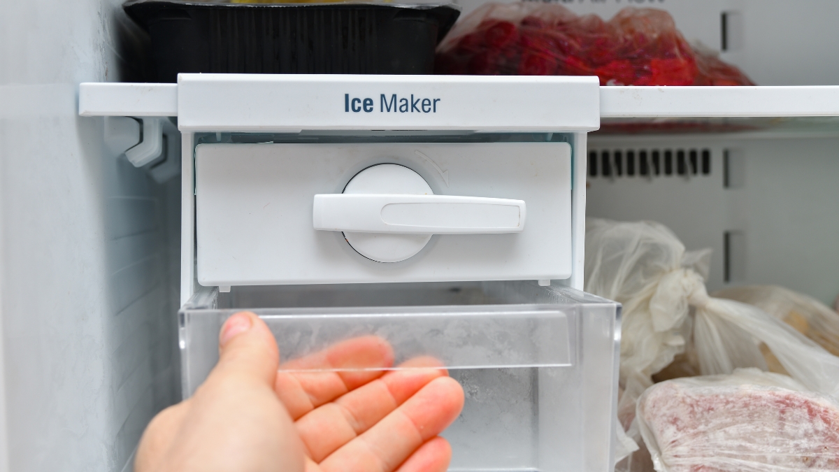 Hand opening a freezer drawer under a home ice maker with frozen food bags around it.
