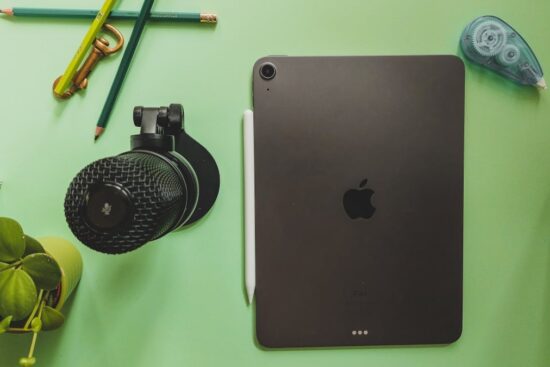 Black Apple iPad Air with Apple Pencil on a green desk beside a microphone, pencils, and small plant.