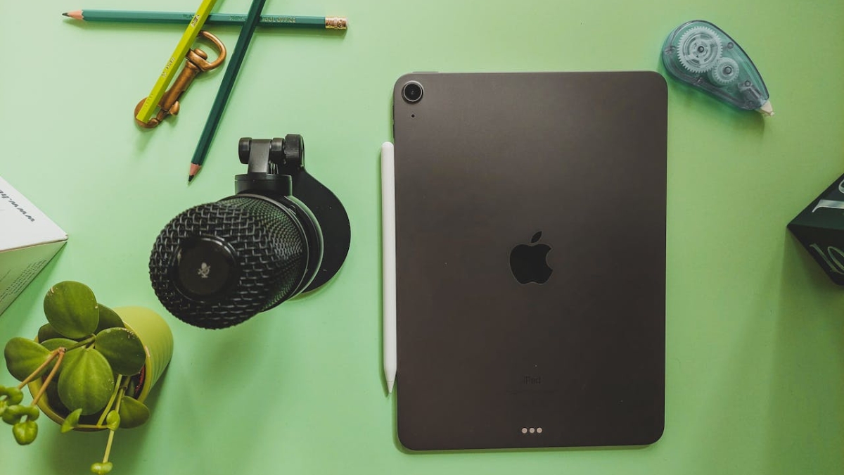 Black Apple iPad Air with Apple Pencil on a green desk beside a microphone, pencils, and small plant.