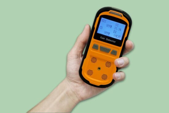 Hand holding an orange 4 gas monitor with a screen showing H2S, CO, O2, and EX gas readings.