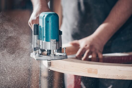 Woodworker using a handheld router to shape the edge of a curved wooden board, with sawdust flying in a well-lit workshop.