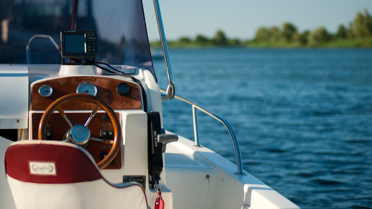 Boat steering console with wooden dash, gauges, and a Lowrance fish finder, set against a calm lake with clear blue skies and distant trees.