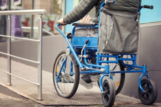 A person in a blue wheelchair going up a metal accessibility ramp outside a building.