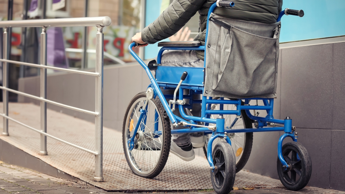 A person in a blue wheelchair going up a metal accessibility ramp outside a building.