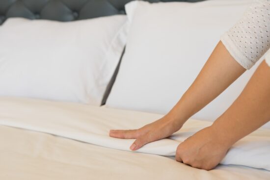 A person smoothing white cotton bed sheets on a neatly made bed with white pillows.