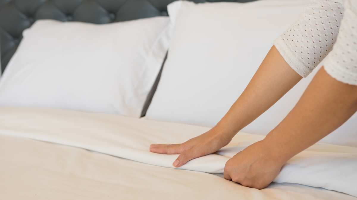 A person smoothing white cotton bed sheets on a neatly made bed with white pillows.