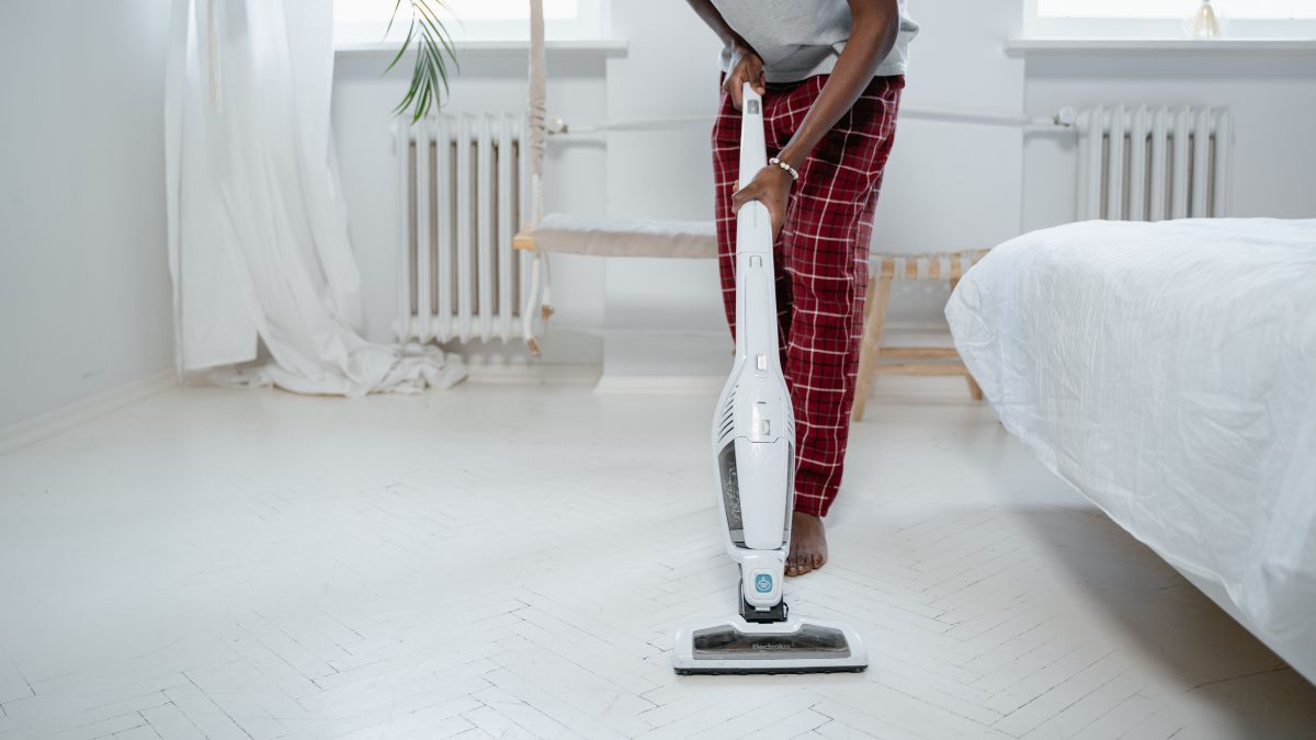 Person cleaning a white bedroom floor with a wet and dry vacuum while wearing red plaid pajama pants.