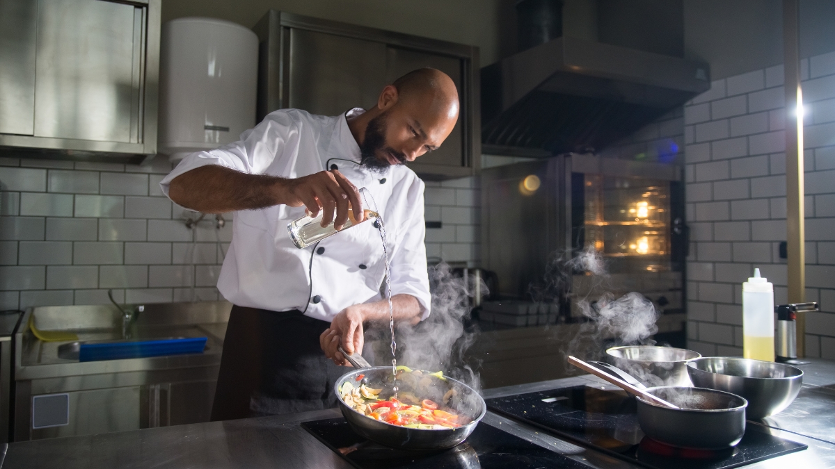 A chef cooking vegetables in a commercial kitchen with steam rising under a canopy hood exhaust fan.