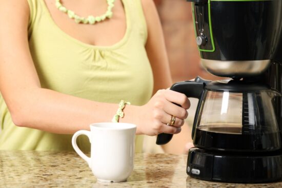 Woman pouring coffee from a drip coffee maker into a white mug on a kitchen counter.