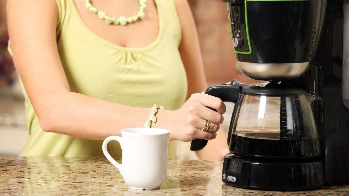 Woman pouring coffee from a drip coffee maker into a white mug on a kitchen counter.