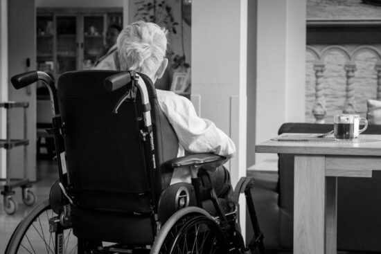 An elderly person in a wheelchair sits at a table with a cup of coffee in a quiet room.
