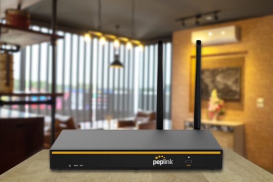 Peplink B-One Gigabit Dual WAN WiFi Router with two antennas on a table in a modern home office.