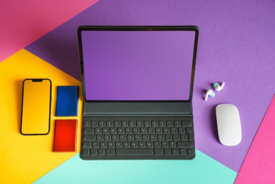 Flat lay of modern tech devices including a tablet with keyboard, smartphone, wireless earbuds, power banks, and a white mouse arranged on a colorful geometric background.