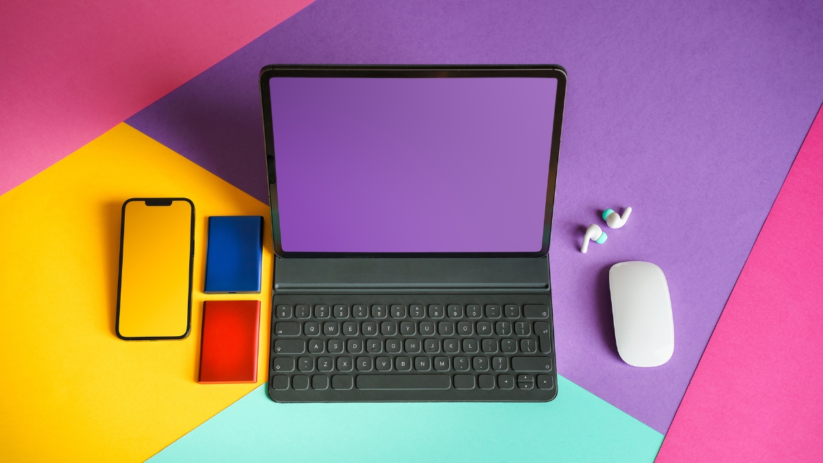 Flat lay of modern tech devices including a tablet with keyboard, smartphone, wireless earbuds, power banks, and a white mouse arranged on a colorful geometric background.