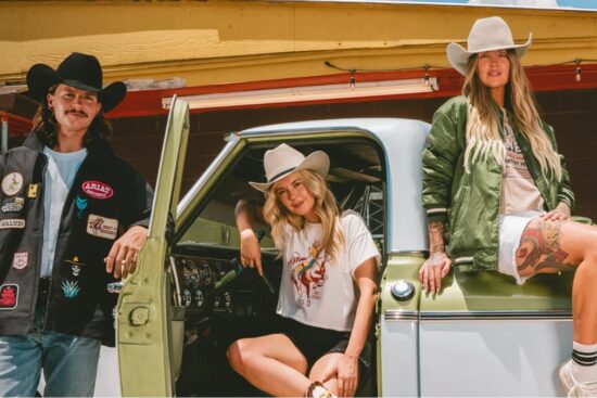 Group of Western-style models wearing Ariat x Sendero apparel and hats while posing with a vintage truck.