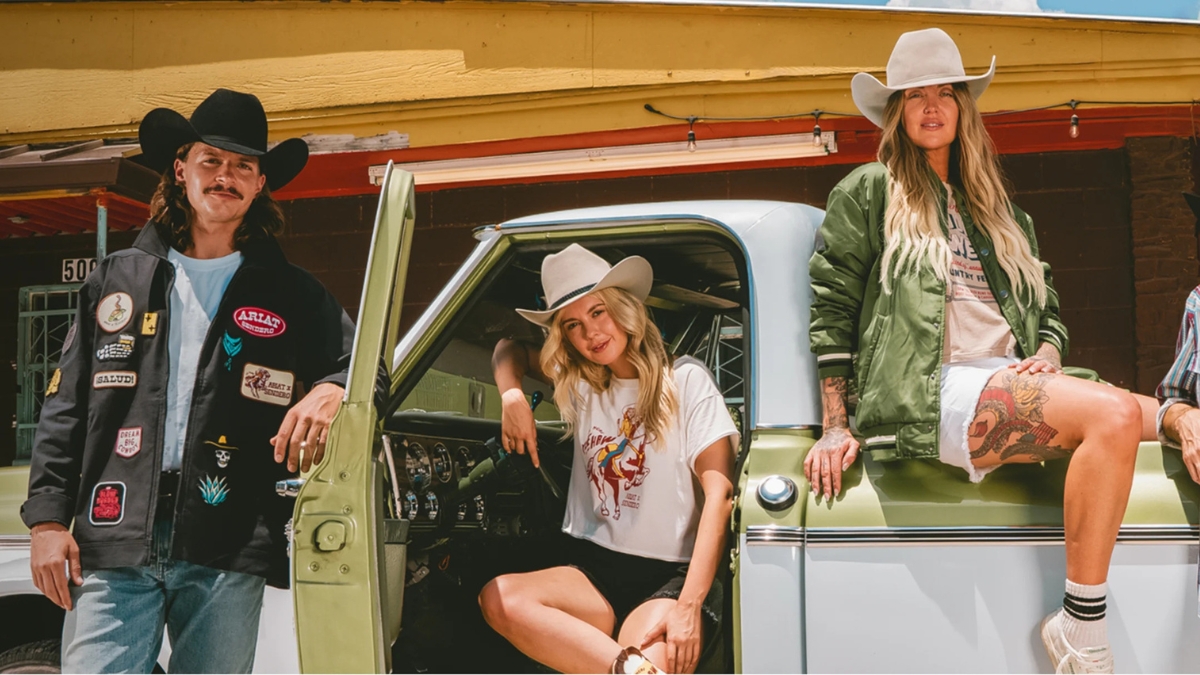 Group of Western-style models wearing Ariat x Sendero apparel and hats while posing with a vintage truck.