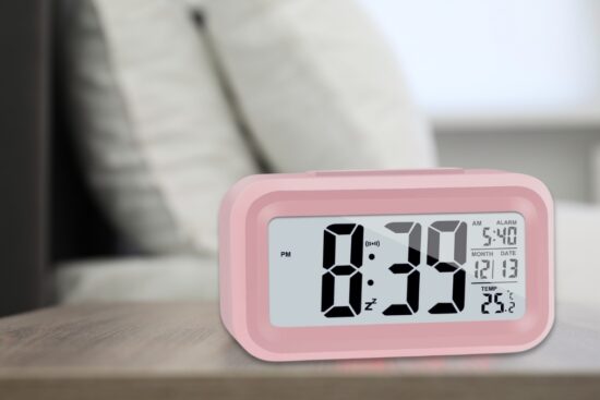 Pink digital alarm clock with large LCD screen showing time, date, alarm setting, and temperature on a bedside table.