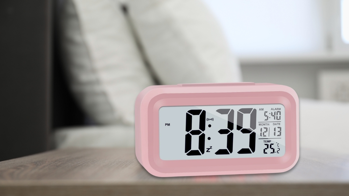 Pink digital alarm clock with large LCD screen showing time, date, alarm setting, and temperature on a bedside table.