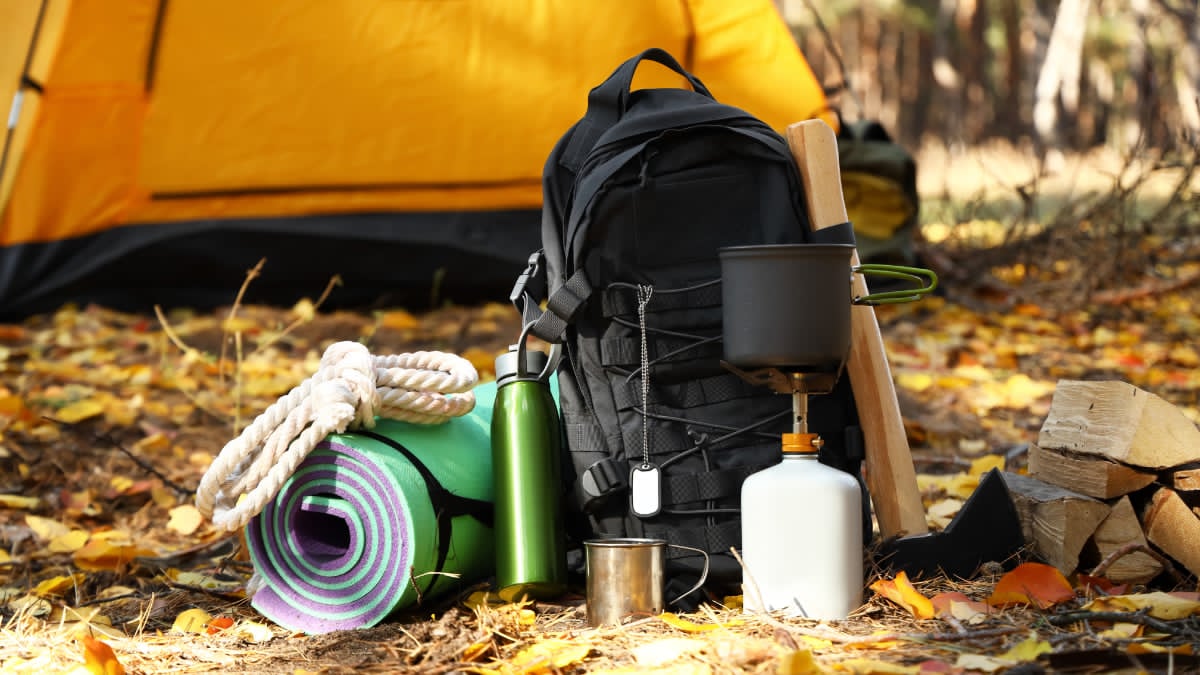 Essential camping gear setup featuring a tent, backpack, sleeping bag, and outdoor cooking supplies at a campsite.